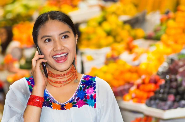 Mooie jonge Spaanse vrouw dragen Andes traditionele blouse met behulp van mobiele telefoon binnen fruitmarkt, kleurrijke gezond voedsel selectie in de achtergrond — Stockfoto