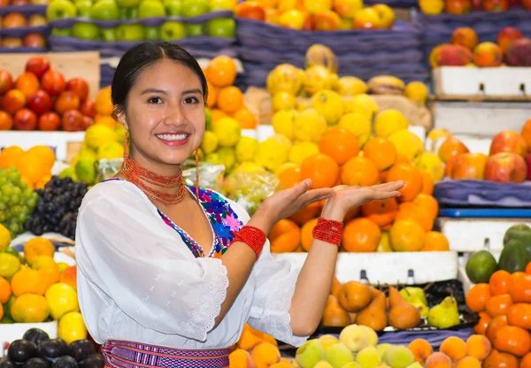 Mooie jonge Spaanse vrouw dragen Andes traditionele blouse poseren houden handen uit voor camera binnen fruitmarkt, kleurrijke gezond voedsel selectie in de achtergrond — Stockfoto