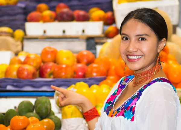 Beautiful young hispanic woman wearing andean traditional blouse pointing using finger inside fruit market, smiling happily, colorful healthy food selection in background