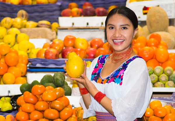 Beautiful young hispanic woman wearing andean traditional blouse posing holding papaya for camera inside fruit market, colorful healthy food selection in background