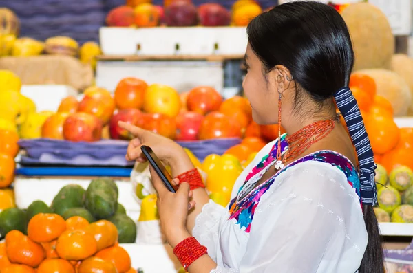 Beautiful young hispanic woman wearing andean traditional blouse pointing using finger, holding mobile phone inside fruit market, colorful healthy food selection in background