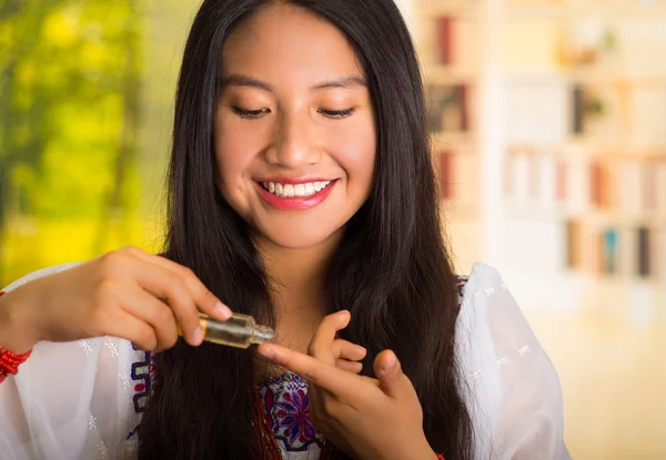 Mulher hispânica bonita vestindo blusa branca com bordado colorido, aplicando produto cosmético ao dedo durante a rotina de maquiagem, sorrindo alegremente, fundo do jardim — Fotografia de Stock