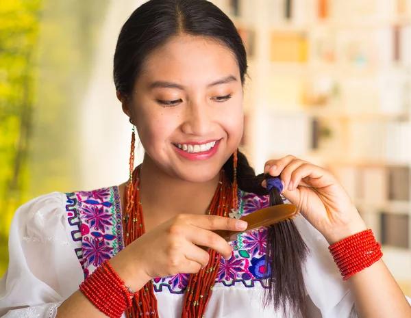 Bella donna ispanica indossa camicetta bianca con ricamo colorato, utilizzando piccola spazzola per capelli durante la routine di trucco, sfondo del giardino — Foto Stock