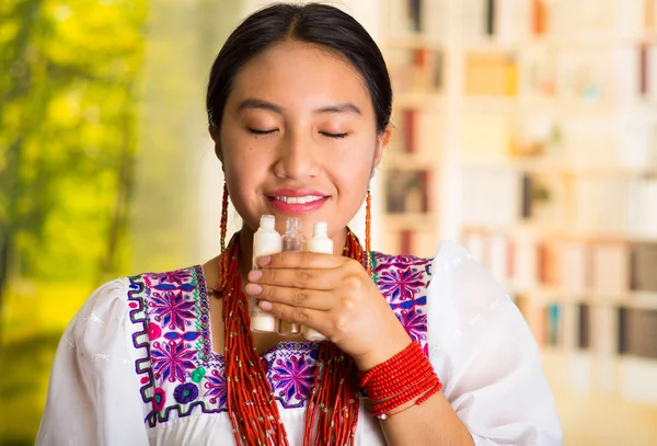 Mulher hispânica bonita vestindo blusa branca com bordado colorido, segurando e cheirando de três pequenos tubos de creme, olhos fechados, durante a rotina de maquiagem, fundo do jardim — Fotografia de Stock