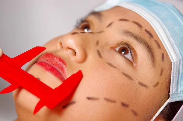 Closeup young womans face preparing for cosmetic surgery with lines drawn on skin, doctor measuring using red tool, as seen from above — Stock Photo, Image