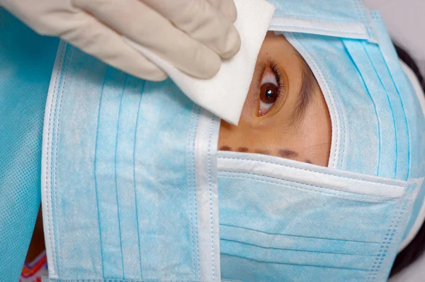 Closeup eye of woman peeking out from total facial cover, preparing for cosmetic surgery concept, doctor wiping with white pad — Stock Photo, Image