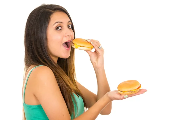Schöne junge Frau posiert vor der Kamera und isst Hamburger, während sie eine andere in der anderen Hand hält, lächelt glücklich, weißer Studiohintergrund — Stockfoto