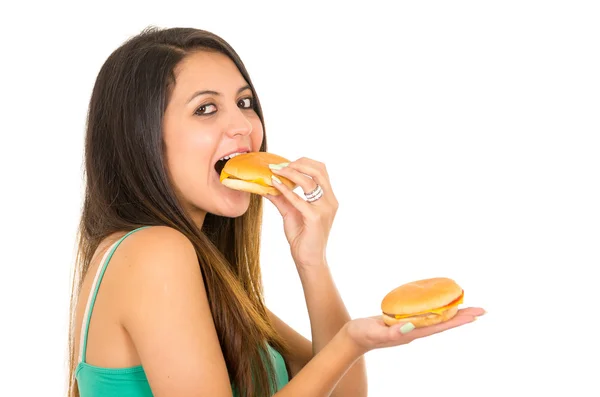 Bella giovane donna in posa per la fotocamera mangiare hamburger mentre ne tiene un altro in altra mano, sorridente felicemente, sfondo bianco studio — Foto Stock