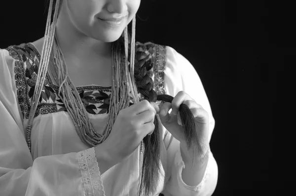 Mulher hispânica jovem bonita vestindo blusa de cor clara com bordas bordadas tradicionais, trançando seu próprio cabelo enquanto sorri, fundo estúdio escuro, edição em preto e branco — Fotografia de Stock