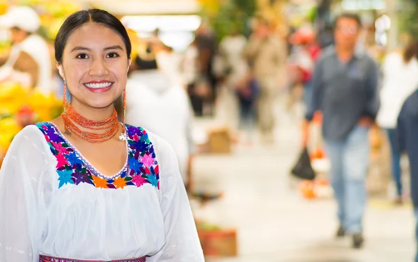 Mooie jonge Spaanse vrouw dragen Andes traditionele blouse poseren voor camera binnen fruitmarkt, kleurrijke gezond voedsel selectie in de achtergrond Stockfoto
