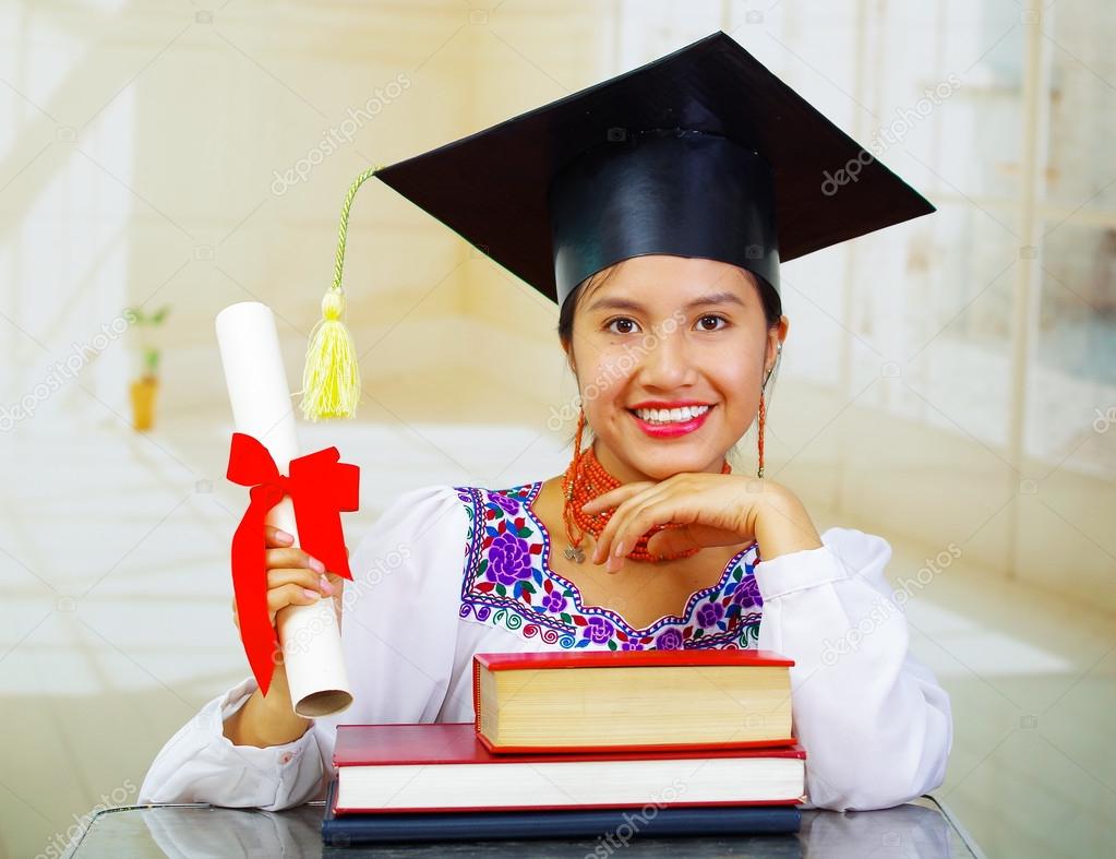 Young female student wearing traditional blouse and graduation hat, sitting by desk with books stacked in front, smiling to camera