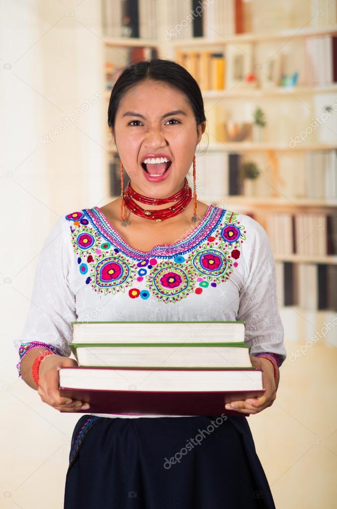 Beautiful young lawyer wearing traditional andean blouse and red necklace, holding stack of books, screaming out to camera, bookshelves background