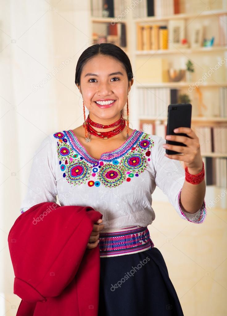 Beautiful young lawyer wearing traditional andean blouse and necklace, holding red jacket while using mobile phone, smiling happily, bookshelves background