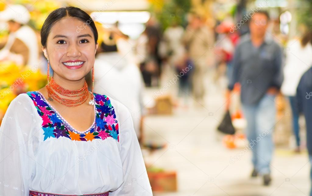 Beautiful young hispanic woman wearing andean traditional blouse posing for camera inside fruit market, colorful healthy food selection in background