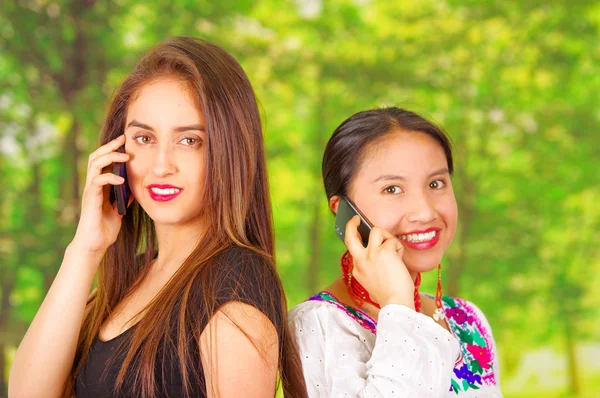 Dos hermosas mujeres jóvenes posando para la cámara, una con ropa tradicional andina, la otra con ropa casual, frotándose las espaldas y ambas hablando en teléfonos móviles, fondo del parque — Foto de Stock