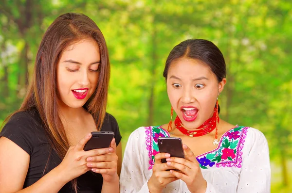 Dos hermosas mujeres jóvenes posando para la cámara, una con ropa tradicional andina, la otra con ropa casual, ambos presionando teléfonos mirando pantallas móviles, fondo del parque — Foto de Stock