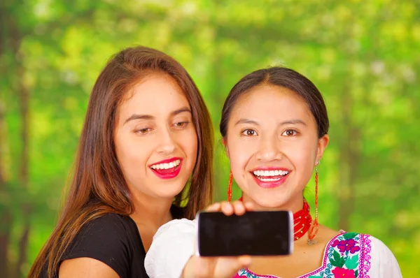 Two beautiful young women standing together facing camera, one wearing traditional andean clothing, the other in casual clothes, holding up mobile posing for selfie smiling, park background — Stock Photo, Image