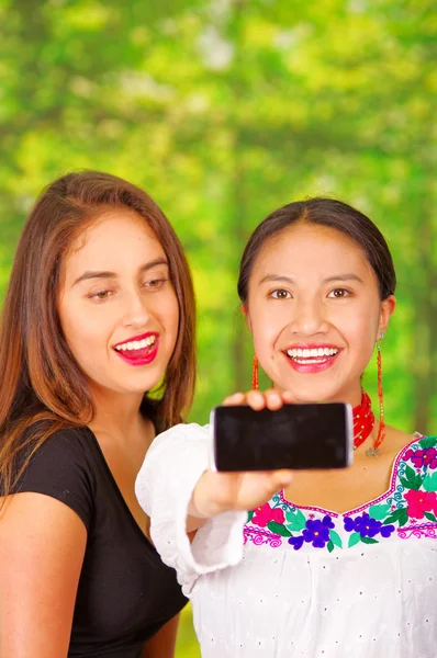 Duas belas mulheres jovens de pé juntos de frente para a câmera, um vestindo roupas tradicionais andinas, o outro em roupas casuais, segurando móvel posando para selfie sorrindo, fundo do parque — Fotografia de Stock