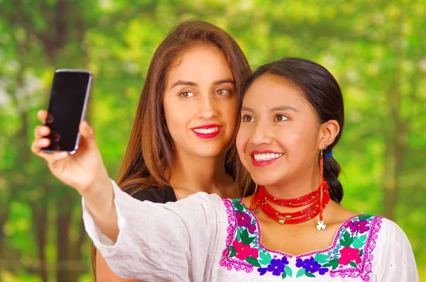 Two beautiful young women standing together facing camera, one wearing traditional andean clothing, the other in casual clothes, holding up mobile posing for selfie smiling, park background — Stock Photo, Image