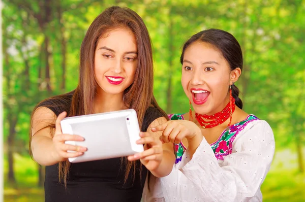 Two beautiful young women posing for camera, one wearing traditional andean clothing, the other in casual clothes, holding tablet between them interacting, both smiling, park background — Stock Photo, Image