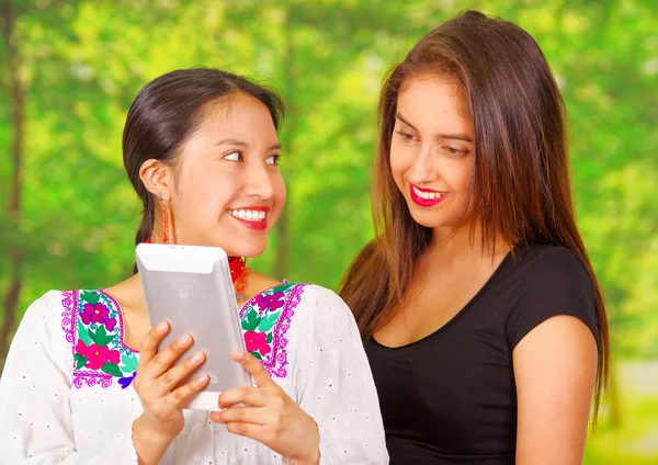 Dos hermosas mujeres jóvenes posando para la cámara, una con ropa tradicional andina, la otra con ropa casual, sosteniendo la tableta entre ellas interactuando, ambas sonrientes, fondo del parque —  Fotos de Stock