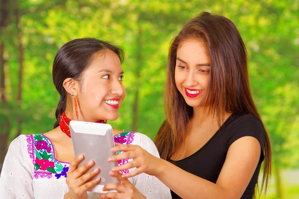 Dos hermosas mujeres jóvenes posando para la cámara, una con ropa tradicional andina, la otra con ropa casual, sosteniendo la tableta entre ellas interactuando, ambas sonrientes, fondo del parque —  Fotos de Stock