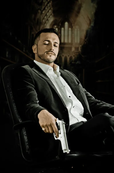 Elegant man sitting in a chair holding  gun over dark background — Stock Photo, Image