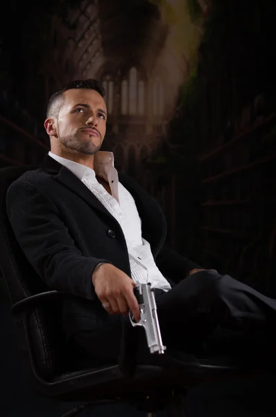 Elegante hombre sentado en una silla sosteniendo pistola sobre fondo oscuro — Foto de Stock