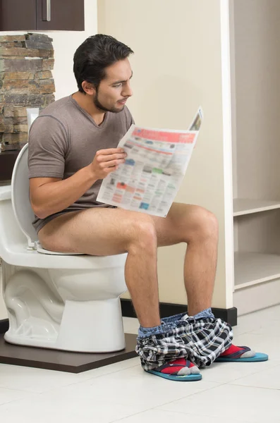 Young relaxed man sitting on the toilet reading newspaper — Stock Photo, Image