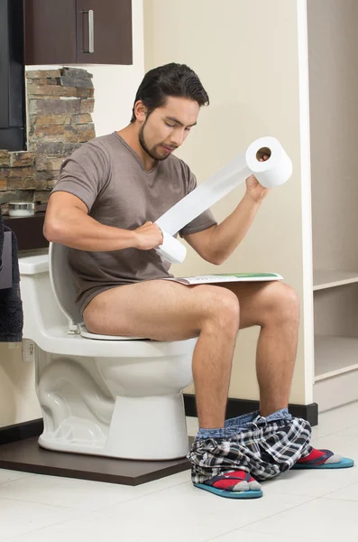 Young relaxed man sitting on the toilet reading newspaper — Stock Photo, Image