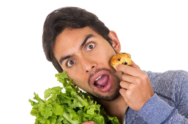 Closeup portrait of confused handsome young man holding fresh lettuce leaves and a muffin — Stock Photo, Image