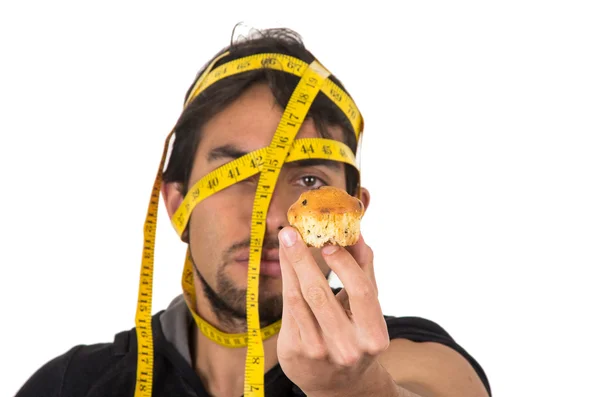 Handsome athletic young man wrapped around measuring tape holding muffin — Stock Photo, Image