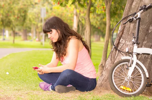 Szép fiatal lány ül a fűben, a park használ sejt telefon — Stock Fotó