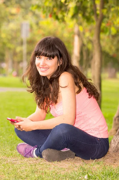 Krásná mladá dívka sedící na trávě v parku pomocí mobilního telefonu — Stock fotografie