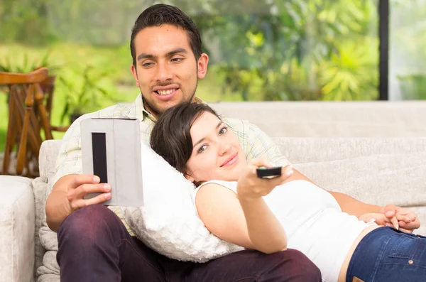 Linda pareja acostada en el sofá viendo la televisión y el uso de la tableta —  Fotos de Stock