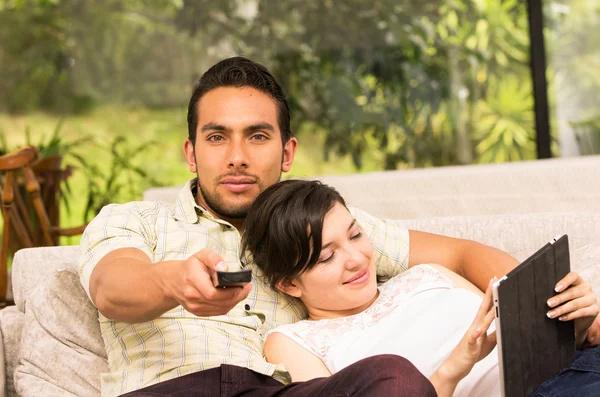 Cute couple cuddling in the sofa while watching tv and using tablet — Stock Photo, Image