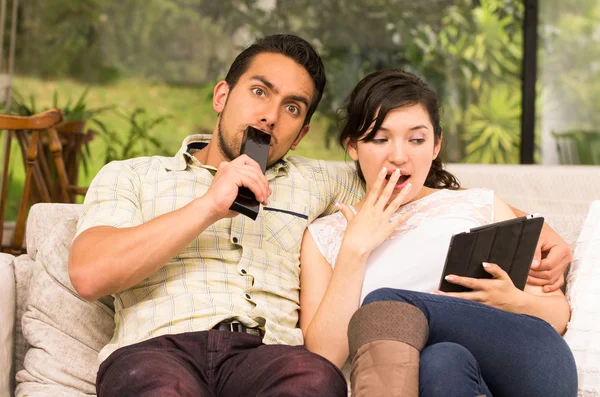 Cute surprised couple cuddling in the sofa while watching tv and using tablet — Stock Photo, Image