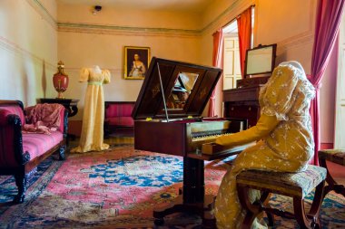 Living room decorated in wooden tables including gold painted details, yellow carpet with a female dress sitting by piano and another attire across simulating daily life at residence Bogota Colombia