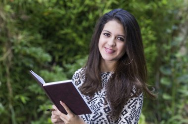 Hispanic brunette business woman in park environment wearing formal clothing holding book open while smiling mouth closed towards camera