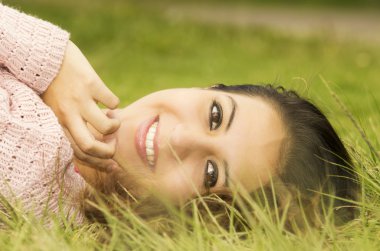 Headshot hispanic female brunette model with head resting on grass and looking sideways towards camera smiling