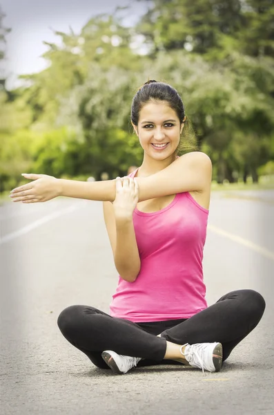 Hispanische Brünette in Yoga-Kleidung sitzt mit überkreuzten Beinen und benutzt den rechten Arm, um den linken zu strecken, während sie in die Kamera lächelt — Stockfoto