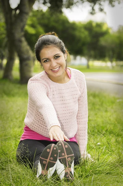 Brune hispanique portant des vêtements de yoga assis sur l'herbe avec les jambes droites en essayant de toucher les pieds en utilisant les bras et en se penchant souriant vers l'avant — Photo