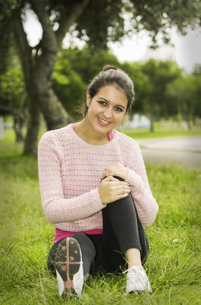 Hispanic brunette sitting on grass in yoga clothing left knee bent with arms wrapped around looking towards camera smiling — Zdjęcie stockowe