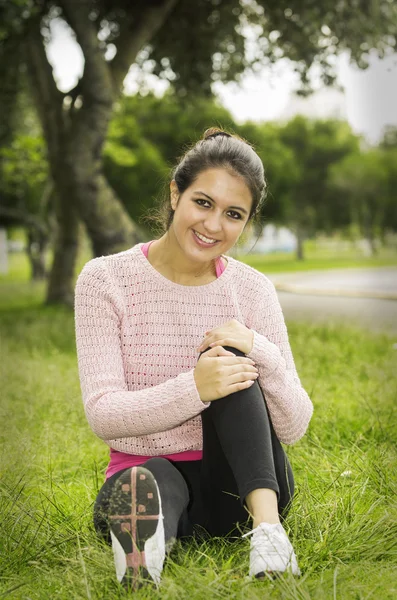 Hispanische Brünette sitzt in Yoga-Kleidung auf dem Gras, das linke Knie gebeugt und die Arme umschlungen und blickt lächelnd in die Kamera — Stockfoto