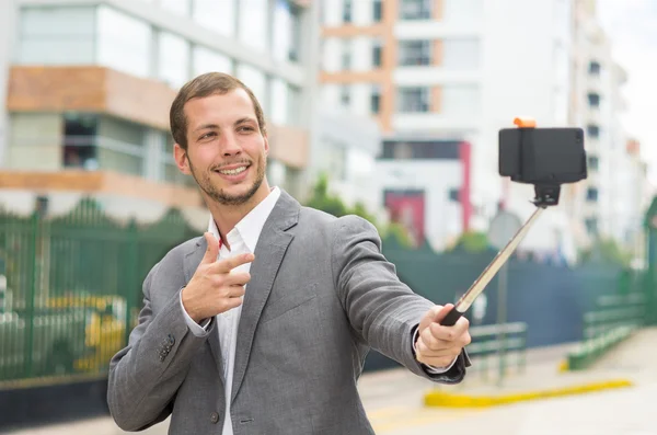 Homme portant des vêtements formels posant avec bâton de selfie en milieu urbain souriant faisant pistolet de main droite — Photo