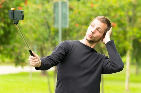 Hispanic man posing with selfie stick in park environment stroking his head using left arm — Stock Photo, Image