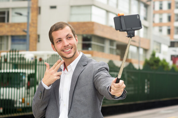 Man wearing formal clothing posing with selfie stick in urban environment smiling using right hand to make a signal
