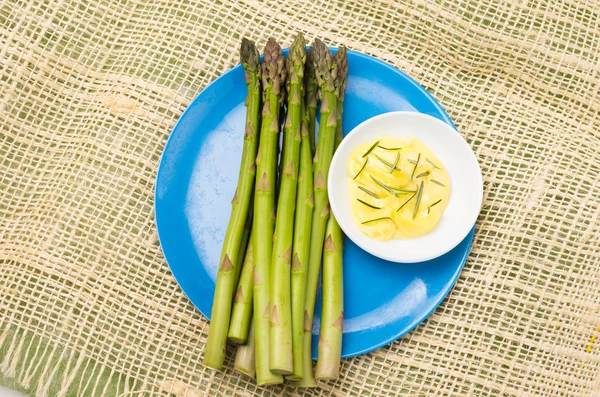 Manojo de espárragos frescos y salsa de mantequilla con romero colocado en un plato azul con mantel de cáñamo . — Foto de Stock