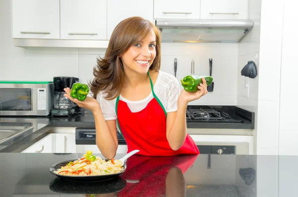 Beautiful woman cooking in modern kitchen posing holding capsicum and smiling with plate of food on counter — Stock Photo, Image