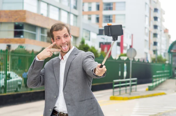 Homem vestindo roupas formais posando com vara selfie em ambiente urbano sorrindo usando a mão direita para fazer um sinal enquanto a língua está fora — Fotografia de Stock